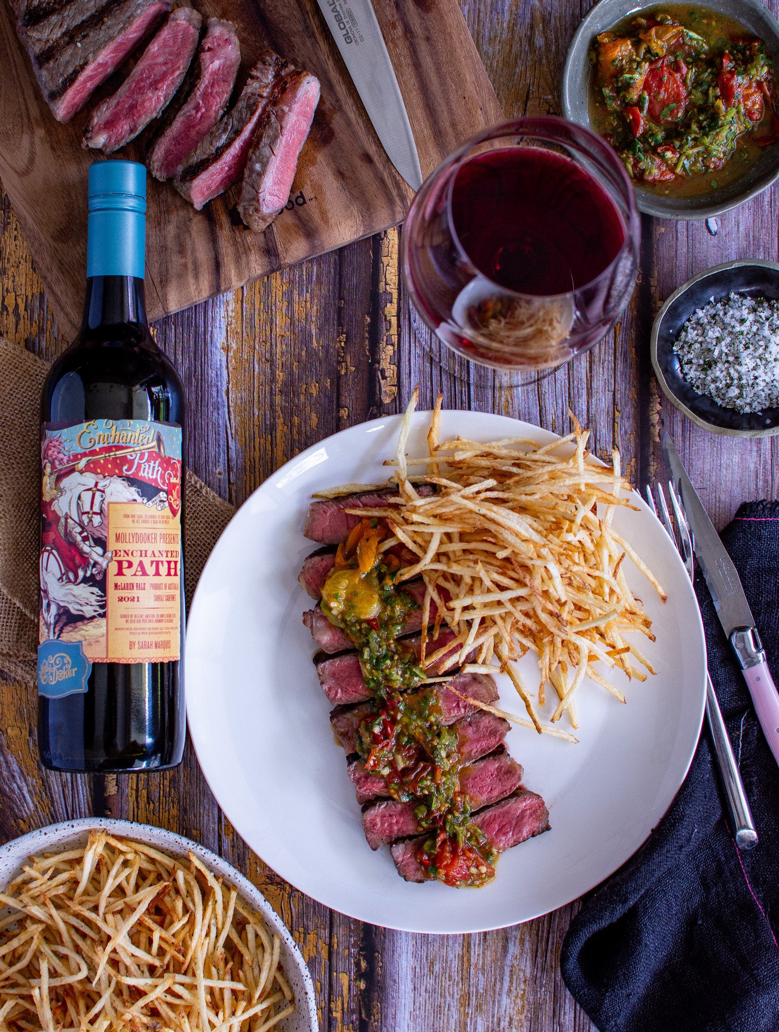 birdseye view of steak with tomato chimichurri on a plate, bottle of mollydooker wine, wine glass and sliced steak on chopping board 