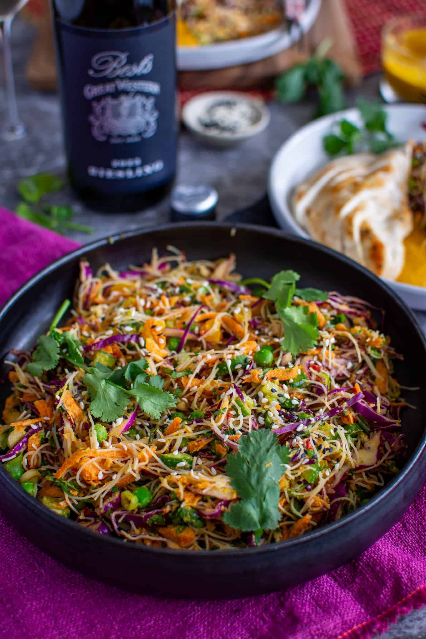 a bowl of cold Asian noodle salad with wine and plates of food in background
