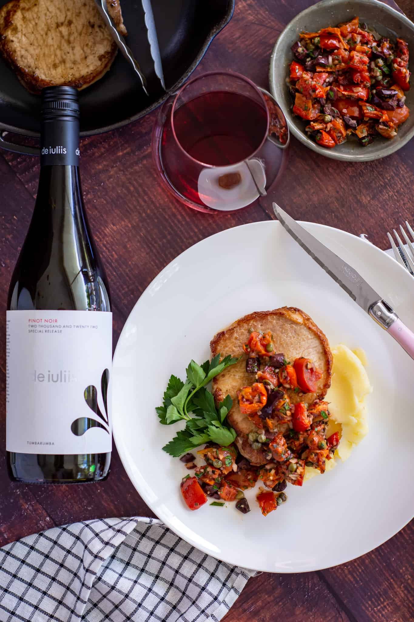 pork chop w/ sundried tomato salsa and mashed potatoes on a white plate. Bottle of red wine next to the plate and a glass of wine too 