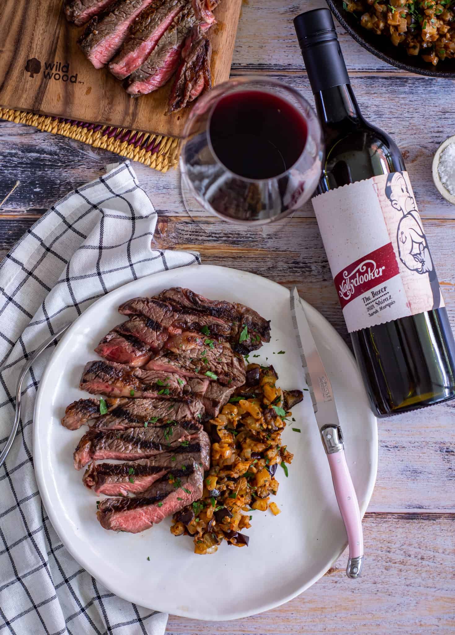 Birdseye view of steak with spiced eggplant on a white plate and a bottle of Mollydooker shiraz next to the plate