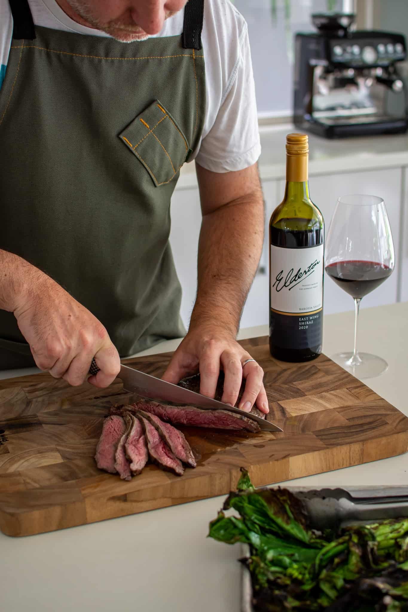 somene slicing steak in the kitchen with red wine beside them
