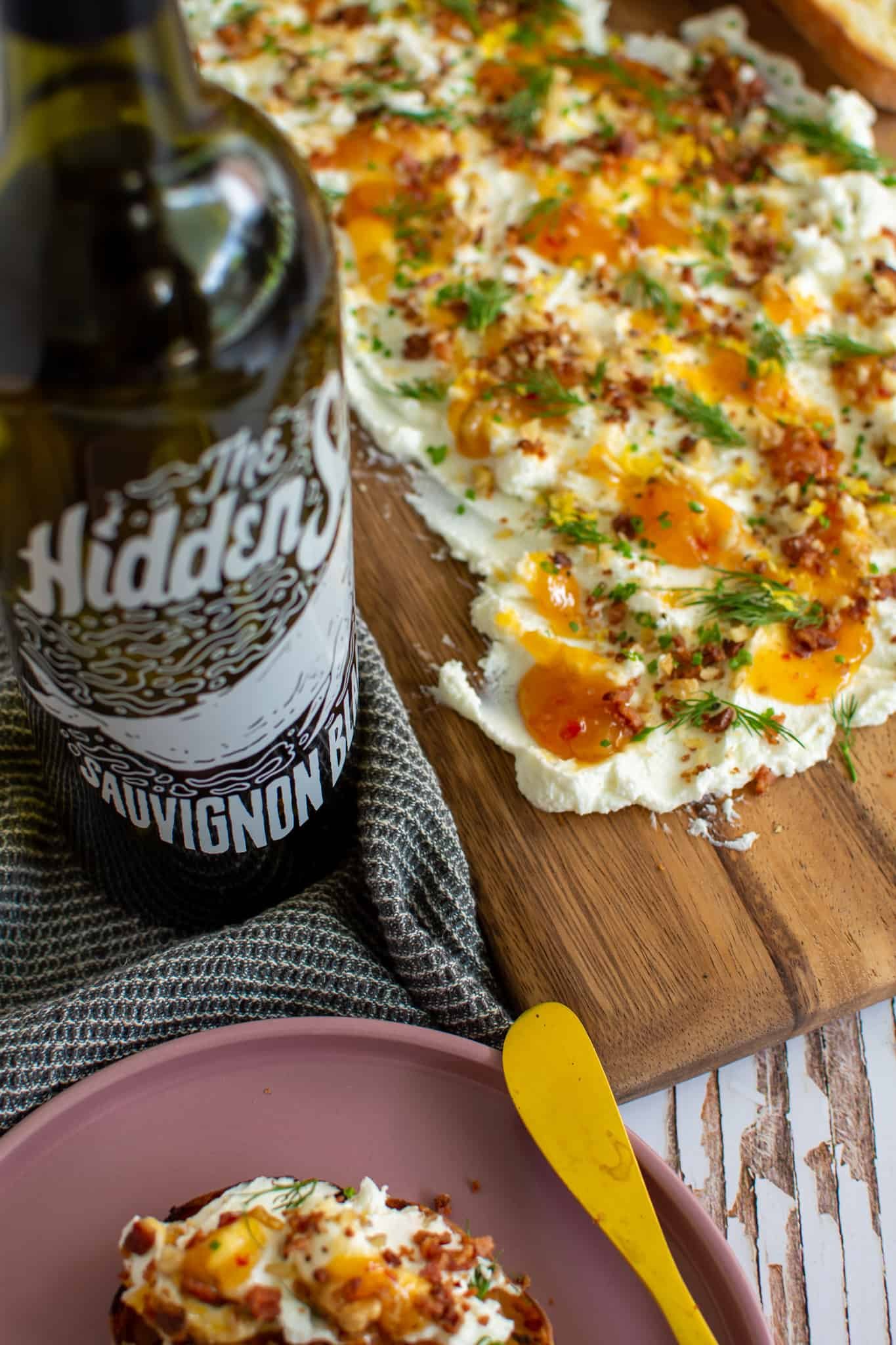 angled photo of goats cheese butter board, bottle of wine and a pink plate with food on toast and golden knife next to it 