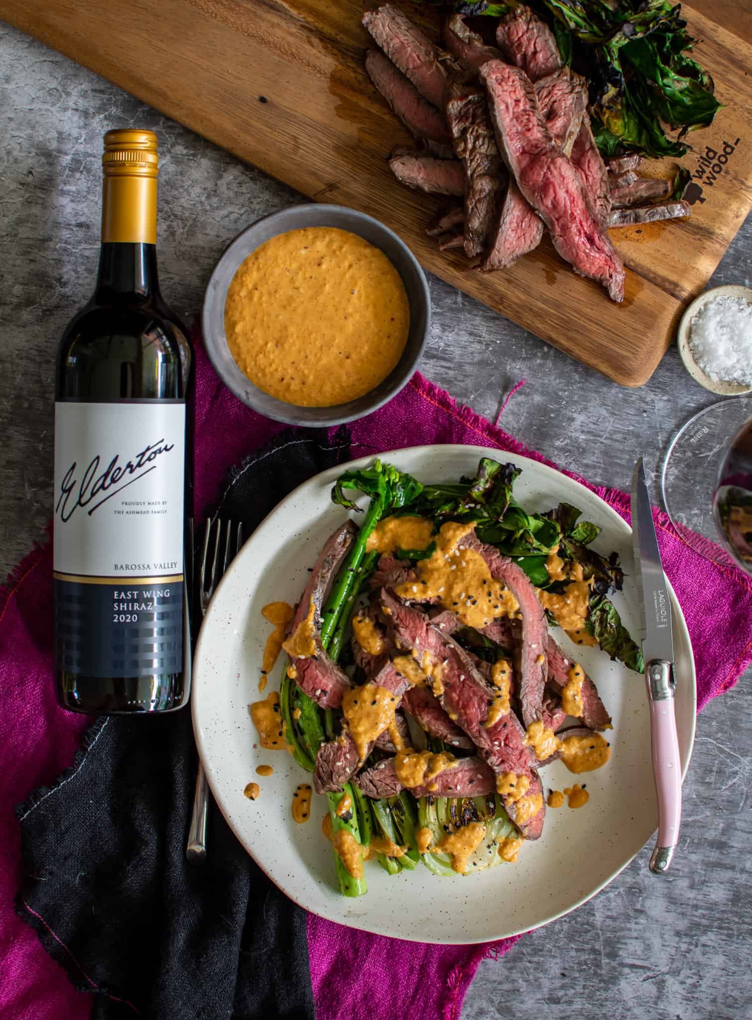 Birdseye view of steak, asian greens and kimchi sauce on a plate with wine lying next to the plate and coloured napkins underneath.