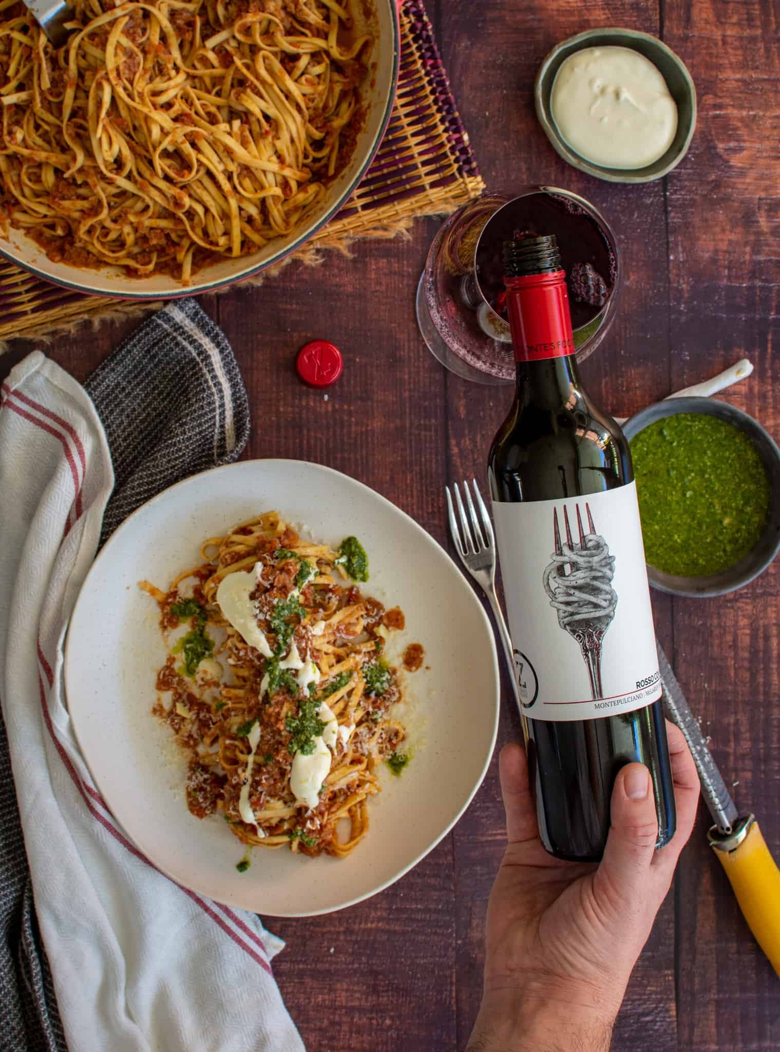 someone pouring wine into a glass with  a bowl of pasta next to it.