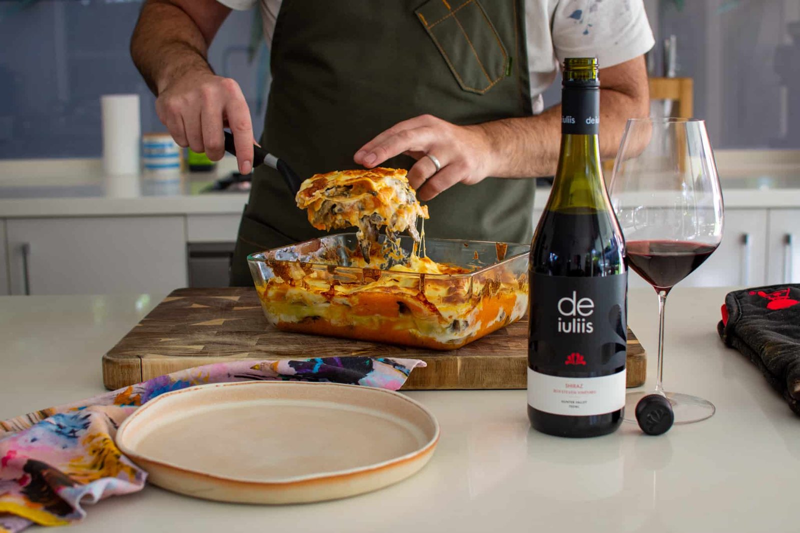 Someone serving vegetable lasagne onto a plate with de iuliis wine in a bottle and glass beside them