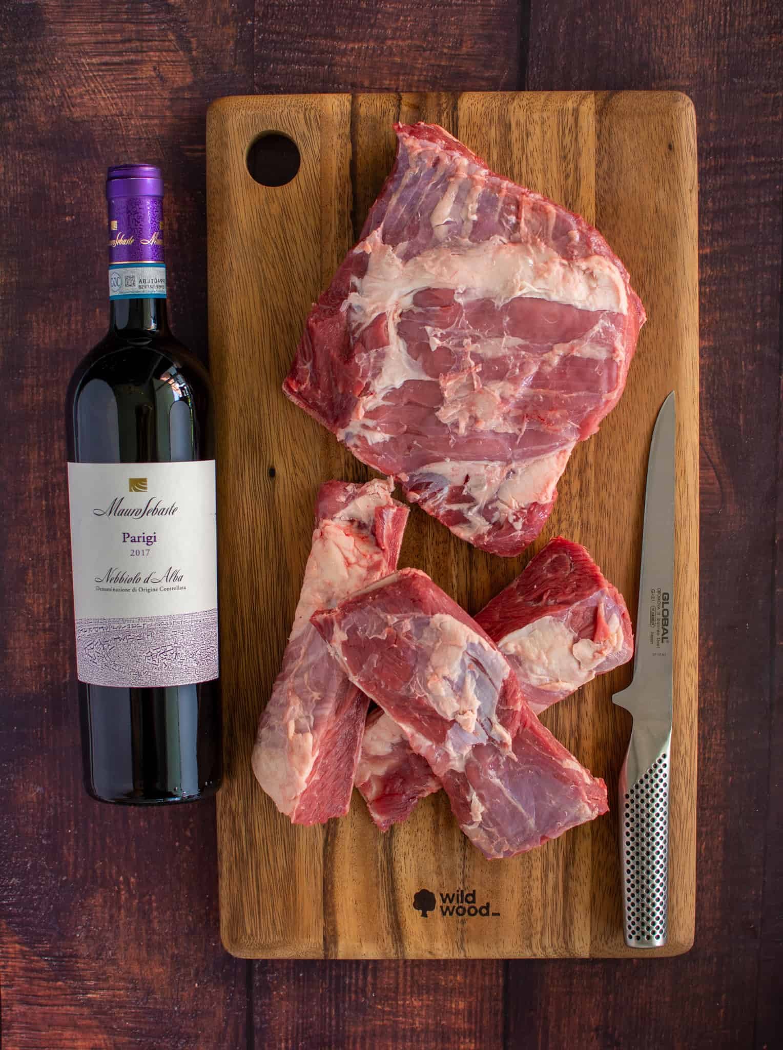 beef short ribs on a chopping board with a knife beside them and a bottle of wine on other side 