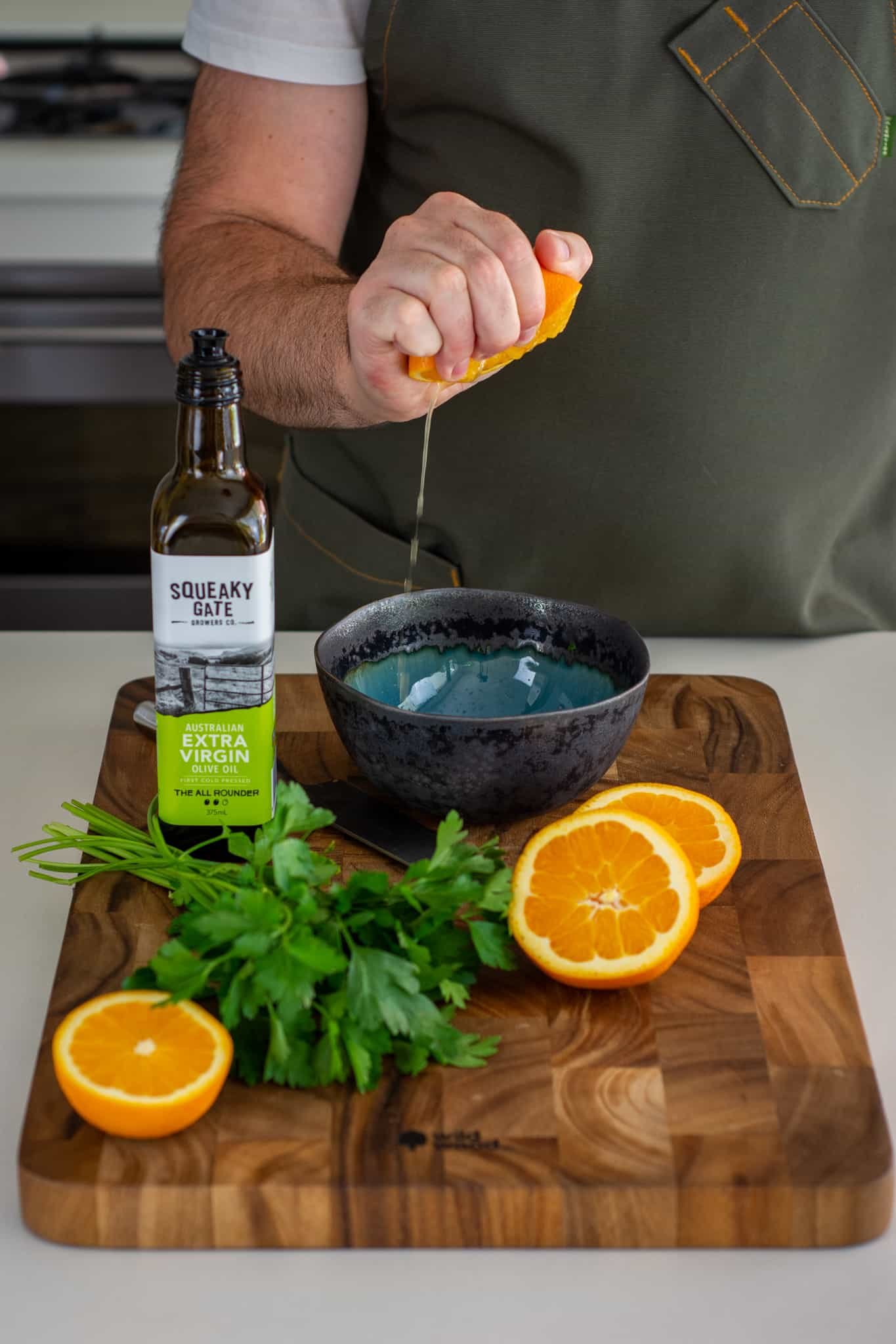 Someone squeezing a orange into a bowl with parsley and olive oil on a chopping board beside the bowl