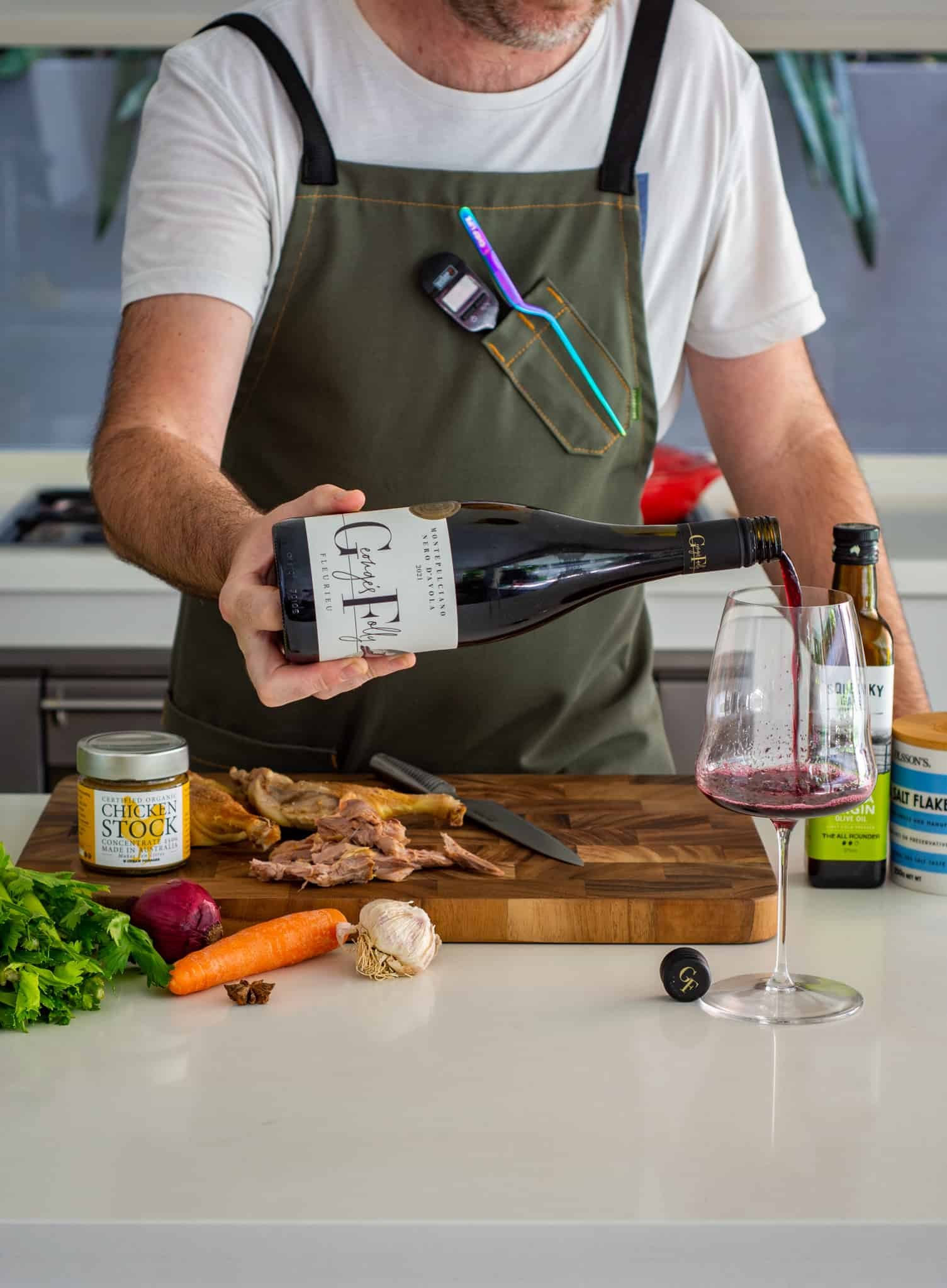 Someone pouing wine in a kitchen with ingredients on a chopping board in front of them 