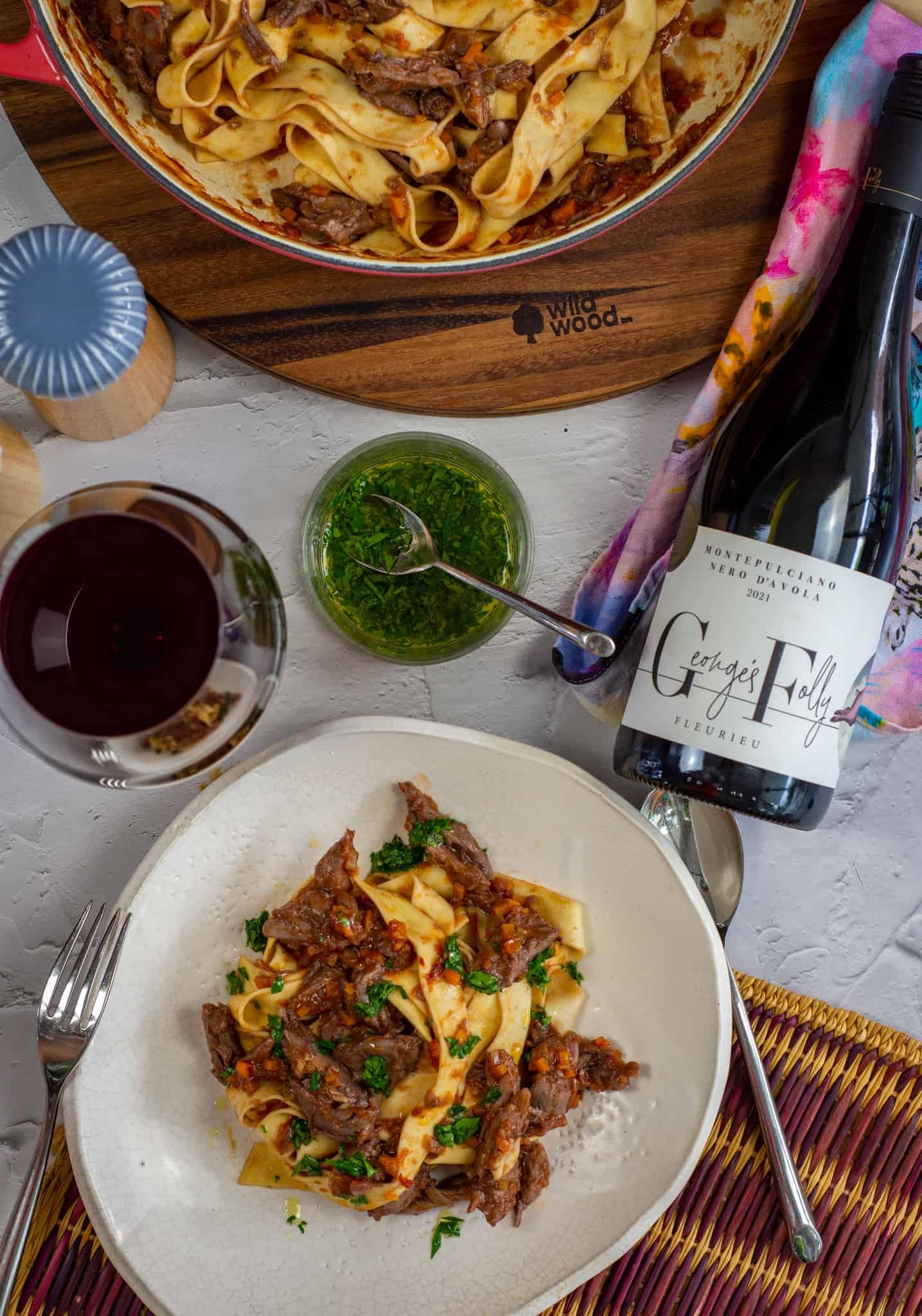 Birdseye view of a plate of duck ragu, wine glass, wine bottle and other items beside it on a table 