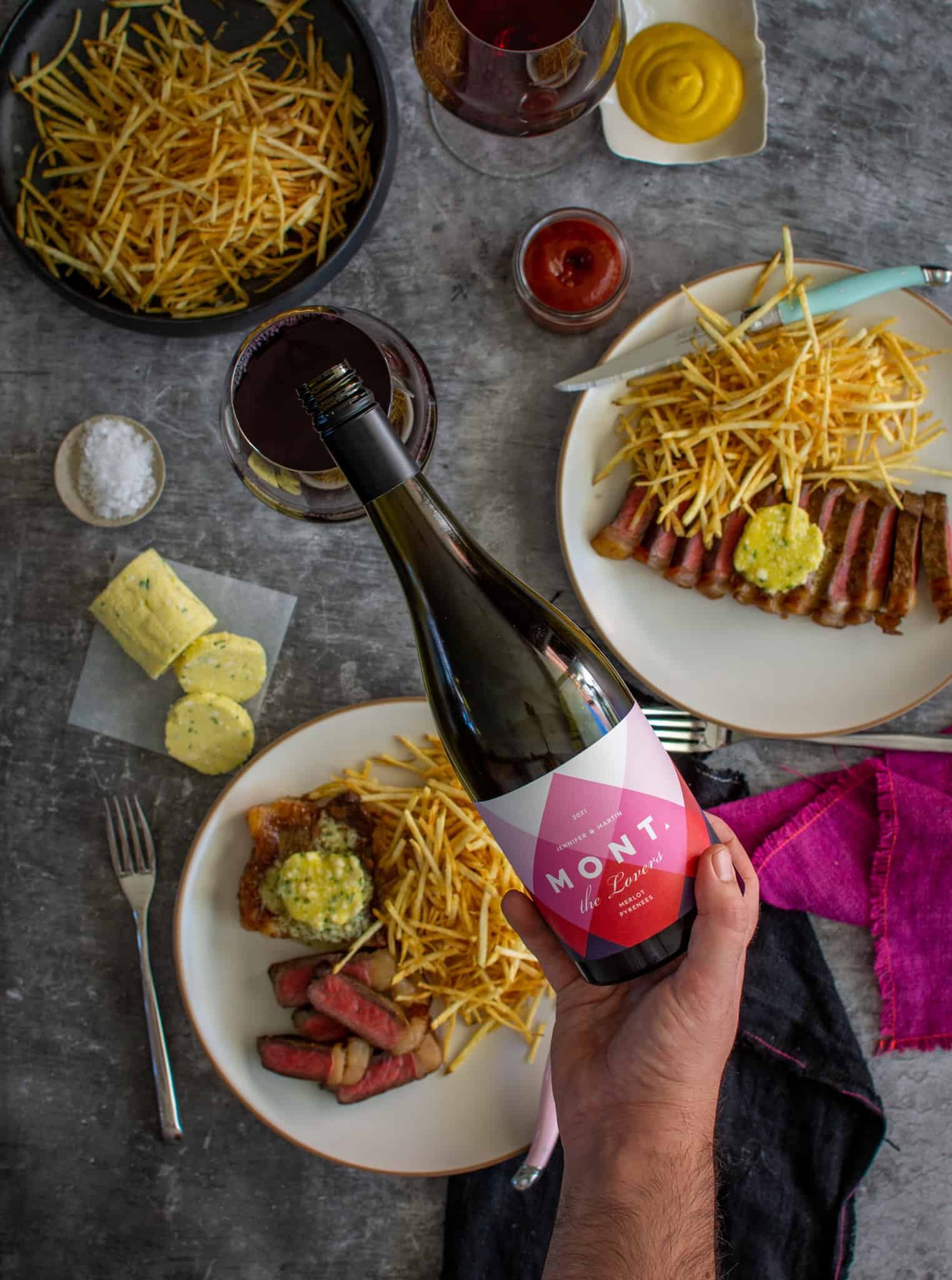 Someone pouring a glass of merlot with striploin steak, frites & blue cheese butter on plates below