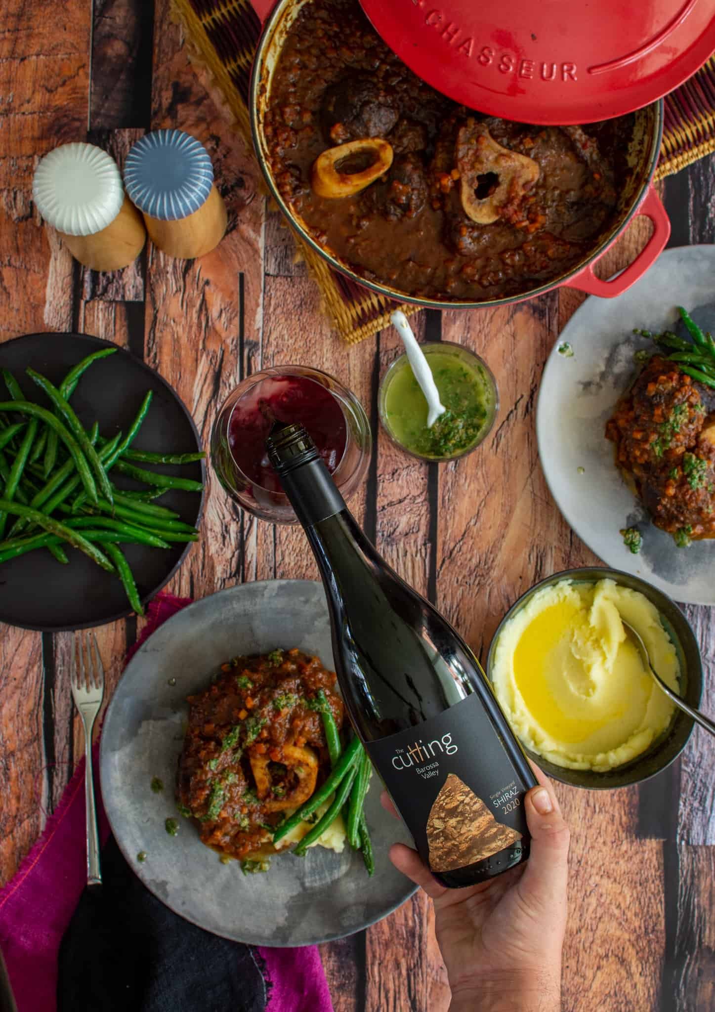 Someone pouring red wine into a glass with plates of beef ossobuco below