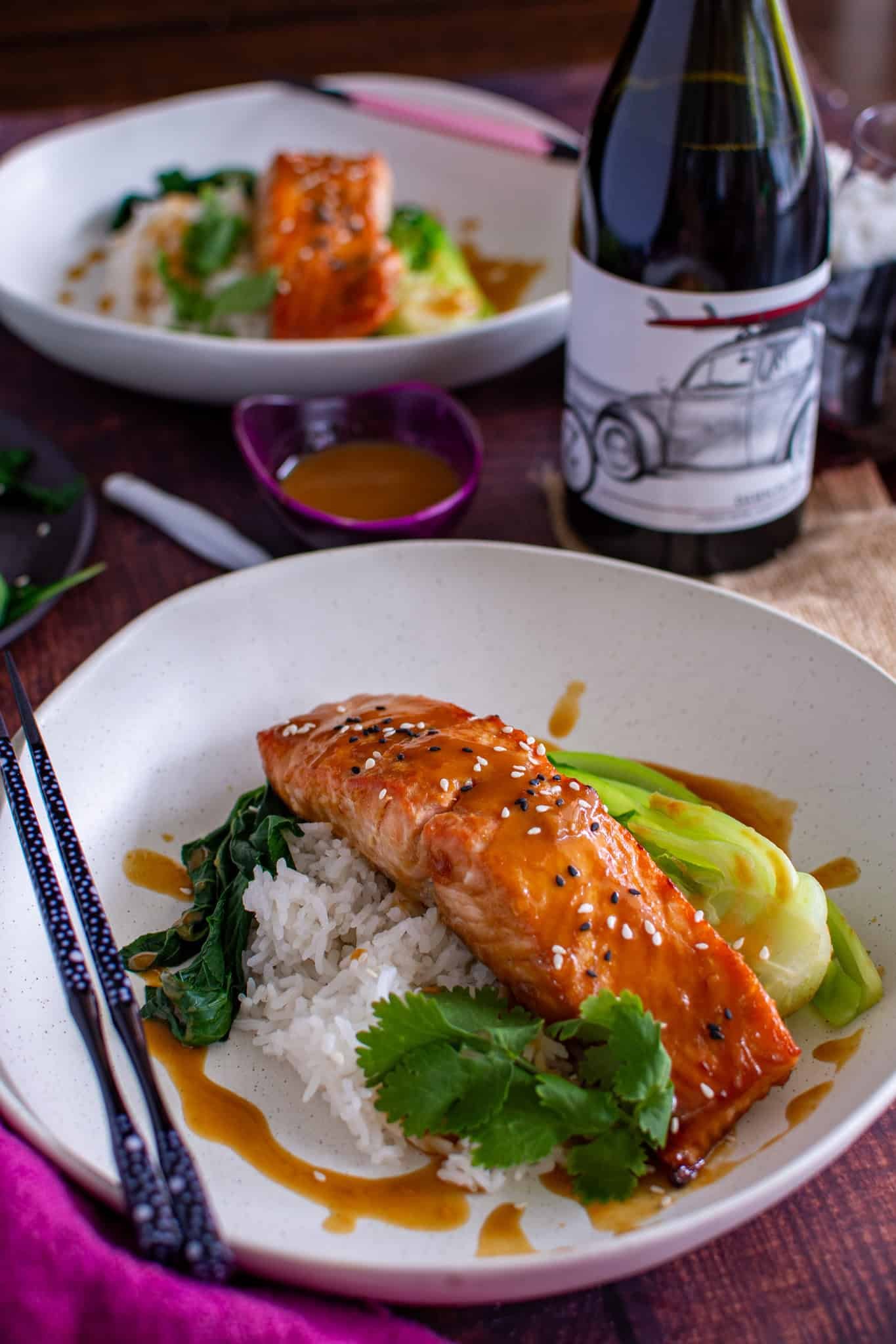 a plate of salmon with rice & asian greens next to a bottle of red wine with another plate in the background