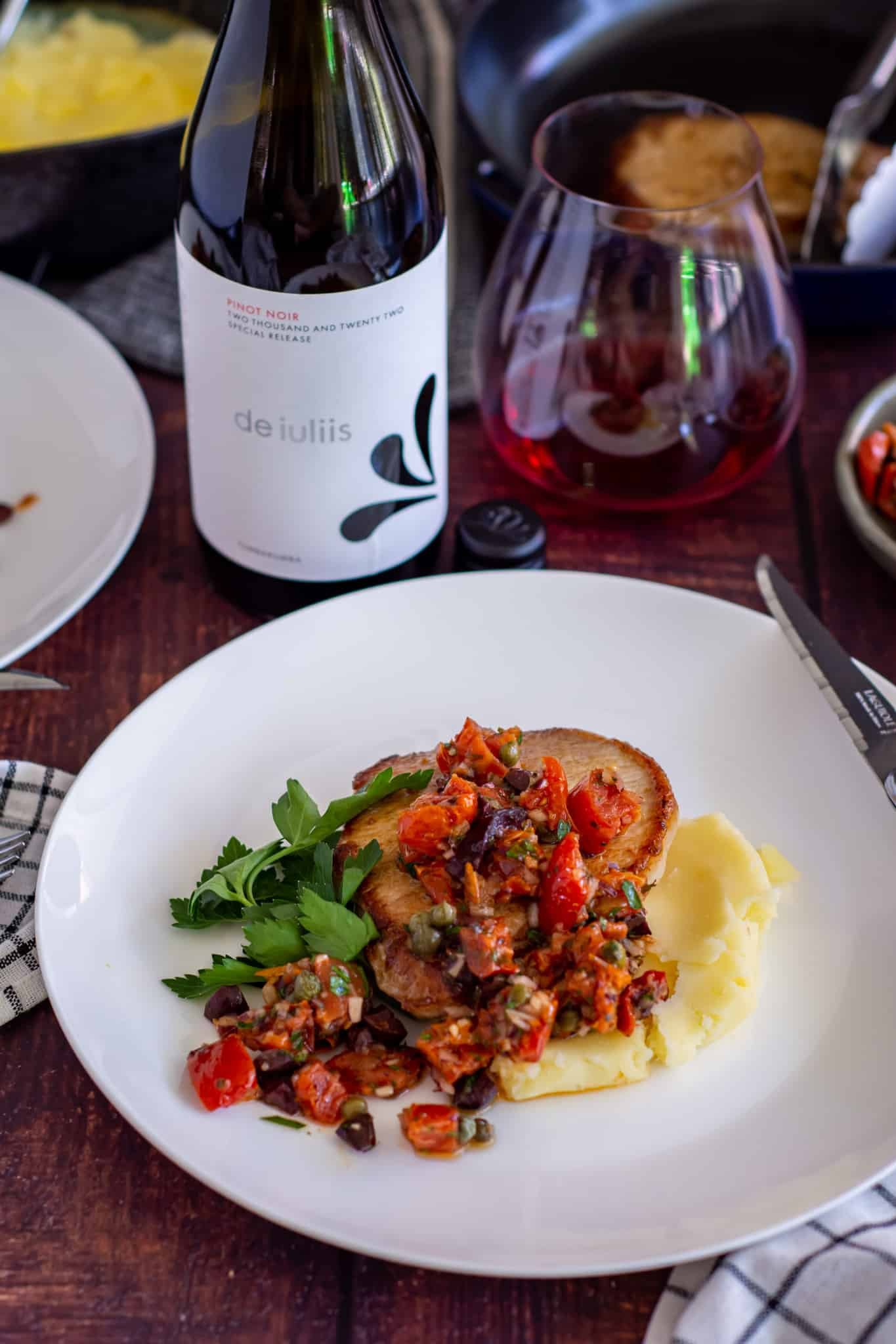 Mediterannean pork chops on a plate with red wine, wine glass and other food in background