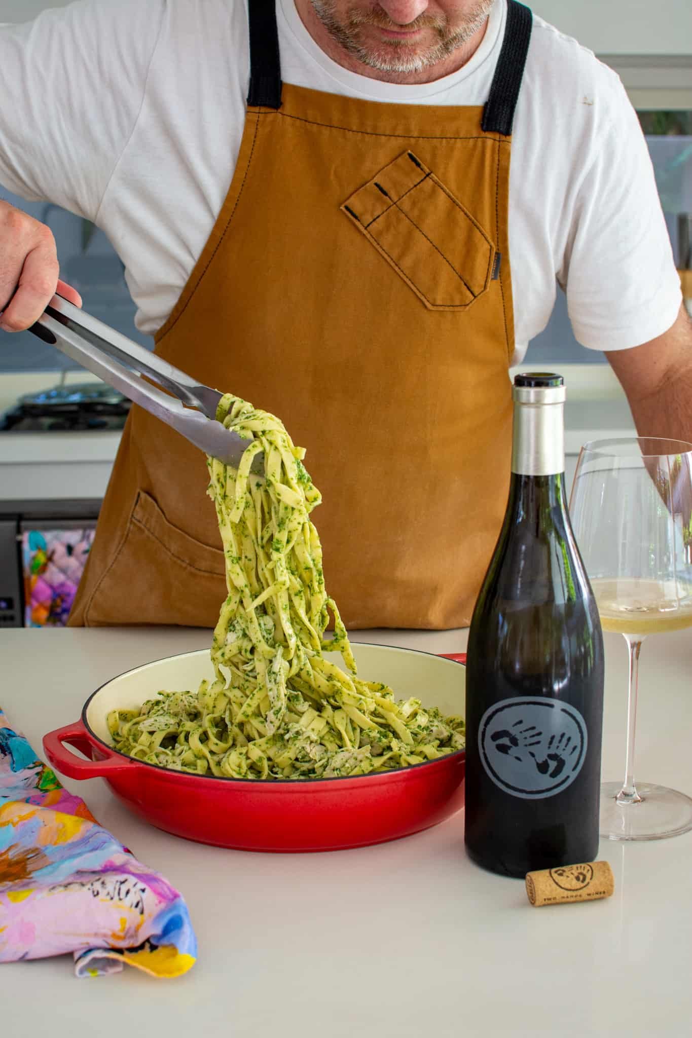 someone picking pasta from a red casserole dish using tongs.  White wine beside them 